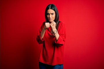 Young beautiful brunette woman wearing casual sweater over isolated red background Ready to fight with fist defense gesture, angry and upset face, afraid of problem