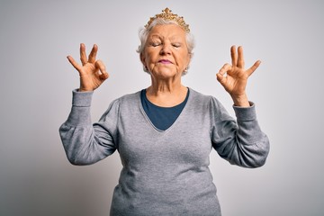 Wall Mural - Senior beautiful grey-haired woman wearing golden queen crown over white background relax and smiling with eyes closed doing meditation gesture with fingers. Yoga concept.