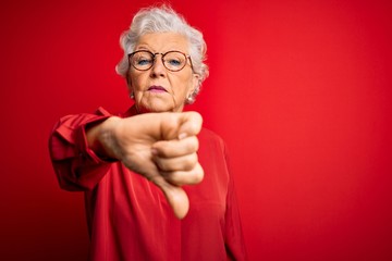 Wall Mural - Senior beautiful grey-haired woman wearing casual shirt and glasses over red background looking unhappy and angry showing rejection and negative with thumbs down gesture. Bad expression.