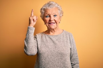 Wall Mural - Senior beautiful woman wearing casual t-shirt standing over isolated yellow background pointing finger up with successful idea. Exited and happy. Number one.