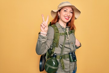 Canvas Print - Young redhead backpacker woman hiking wearing backpack and hat over yellow background smiling looking to the camera showing fingers doing victory sign. Number two.