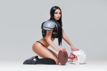 Wall Mural - Side view of beautiful sexy brunette dressed in bikini and American football uniforms posing sitting on the floor with a ball and helmet isolated on grey background