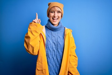 Sticker - Young blonde woman with short hair wearing rain coat for rainy weather over blue background pointing finger up with successful idea. Exited and happy. Number one.