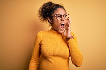 Sticker - Young beautiful african american girl wearing sweater and glasses over yellow background shouting and screaming loud to side with hand on mouth. Communication concept.