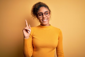 Sticker - Young beautiful african american girl wearing sweater and glasses over yellow background pointing finger up with successful idea. Exited and happy. Number one.