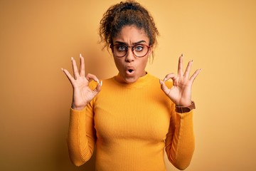 Young beautiful african american girl wearing sweater and glasses over yellow background looking surprised and shocked doing ok approval symbol with fingers. Crazy expression