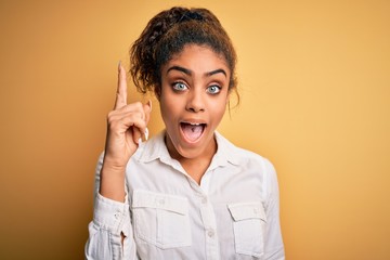 Sticker - Young beautiful african american girl wearing casual shirt standing over yellow background pointing finger up with successful idea. Exited and happy. Number one.