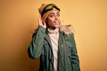 Wall Mural - Young african american afro skier girl wearing snow sportswear and ski goggles smiling with hand over ear listening an hearing to rumor or gossip. Deafness concept.