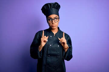 Sticker - Young african american chef girl wearing cooker uniform and hat over purple background Pointing up looking sad and upset, indicating direction with fingers, unhappy and depressed.