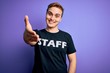 © Krakenimages.com - Young handsome redhead worker man wearing staff t-shirt uniform over purple background smiling friendly offering handshake as greeting and welcoming. Successful business.