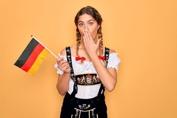 Wall Mural - Beautiful blonde patriotic woman with blue eyes wearing octoberfest dress holding german flag cover mouth with hand shocked with shame for mistake, expression of fear, scared in silence, secret