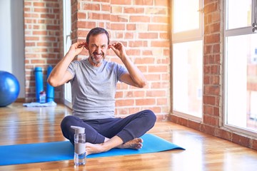 Poster - Middle age handsome sportman sitting on mat doing stretching yoga exercise at gym Smiling pulling ears with fingers, funny gesture. Audition problem