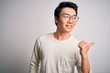 © Krakenimages.com - Young handsome chinese man wearing casual t-shirt and glasses over white background smiling with happy face looking and pointing to the side with thumb up.