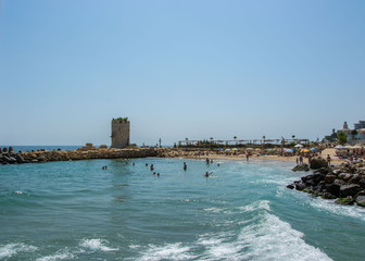 Seascape, view of a beach full with people, old tower of a castle and rocks and tetrapods, blue water and sky, summer season, nature