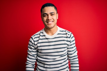 Wall Mural - Young brazilian man wearing casual striped t-shirt standing over isolated red background with a happy and cool smile on face. Lucky person.