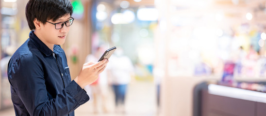 Smart Asian man wearing glasses using smartphone in shopping mall or department store. Telecommunication network and technology. Mobile phone app for urban lifestyle. 