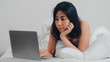 © tirachard - Young Asian woman using laptop checking social media feeling happy smiling while lying on bed after wake up at house in the morning, Attractive thai girl smiling relax in bedroom at home concept.