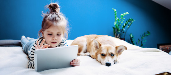 Cute girl watching cartoons with little dog at home using wireless tablet computer. Toddler growing with gadget in cozy place