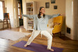 © shurkin_son - Full length shot of happy energetic mature woman in casual clothes exercising at home because of social distancing, practicing yoga on mat, standing in warrior ii pose. Age, wellness and health