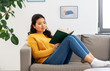 © Syda Productions - people and leisure concept - happy asian young woman in yellow sweater sitting on sofa and reading book at home
