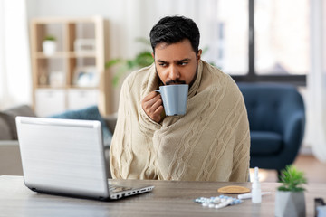 health, cold and people concept - sick young indian man in blanket with laptop computer drinking hot tea and working at home