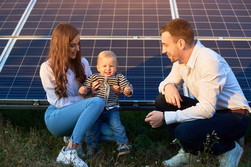 Wall Mural - Young family of three is crouching near photovoltaic solar panel, little boy is looking at camera, parents looking at him, modern family concept