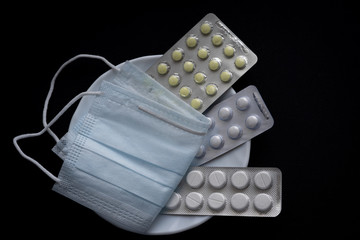 White tablets in blister and blue medical face mask on white plate isolated on dark black background