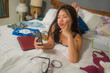slackness and disorganized during covid-19 home lockdown - young disorderly and chaotic Asian Chinese woman on bed using internet mobile phone on grimy messy bedroom