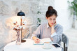 © Marina April - Young woman writing in notebook and reading from mobile phone in cozy cafe indoors. Freelancer working remotely from home office with table lamp and coffee. Comfortable coworking modern workplace.