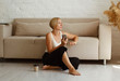 © Sunny - Young beautiful woman sitting on floor near cosy sofa with cup of coffee or tea searching in mobile