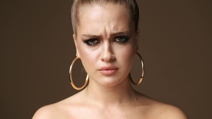 Poster - A displeased young half-naked woman wearing stylish earrings is looking angry and offended isolated over beige background in studio