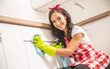 Lovely young housemaid in apron cleaning white kitchen cupboard door with a cloth and detergent in yellow rubber gloves
