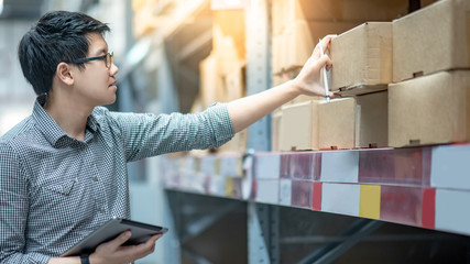 Wall Mural - Young Asian man worker doing stocktaking of product in cardboard box on shelves in warehouse by using digital tablet and pen. Physical inventory count concept