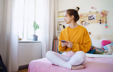 Poster - Sad young female student sitting on bed, using smartphone.