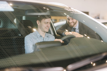 Wall Mural - Happy young man behind the wheel of new car in auto dealership. Professional car salesman wearing business suit is telling about new car model. Concept of choosing and buying new car at showroom.