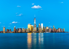 Illuminated One World Trade Center And Buildings In City By Hudson River Against Blue Sky