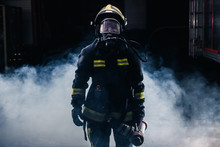 Portrait Of A Female Firefighter Wearing A Helmet And All Safety Equipment A While Holding A Tomahawk And Wearing An Oxygen Mask Indoors Surrounded By Smoke