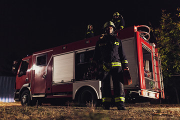 Wall Mural - Professional firefighter standing in uniform wearing protective helmet and holding water hose. Firetruck in the background.