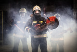 Fototapeta  - Group of professional firefighters wearing full equipment, oxygen masks, and emergency rescue tools, circular hydraulic and gas saw, axe, and sledge hammer. smoke and fire trucks in the background.