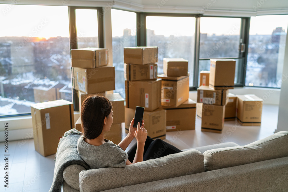 Home move out of apartment moving boxes woman using online movers services on mobile phone app easy pick-up with packages for new home. Asian new homeowner girl happy sitting in sofa. - obrazy, fototapety, plakaty 