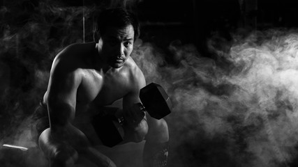 Wall Mural - asian athletic strong man sitting and having weight lifting with dumbbells with one arm in gym and fitness center with smoke and dark background in black and white