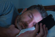 © TheVisualsYouNeed - attractive man falling asleep on bed late at night in dark light while networking on mobile phone or using online dating in internet and social media app addiction concept