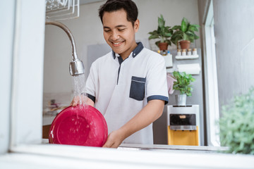 Wall Mural - Happy young asian man in white shirt standing and washing dishes on the kitchen