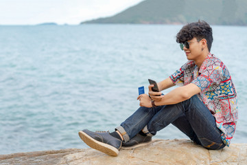 Wall Mural - Smiling young smart Asian man guy sitting on rocky seacoast beach using smartphone with internet for online shopping and credit card for e-banking payment. E-commerce and wireless technology concept