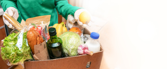 Woman in protective gloves taking food products out of delivered box in kitchen. Safe delivery. Banner, copy space.