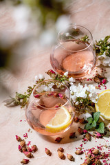 Wall Mural - Cocktail glasses of pink rose champagne, cider or lemonade with dry rose buds, lemon and mint. Blossom cherry branches above. Pink marble background.