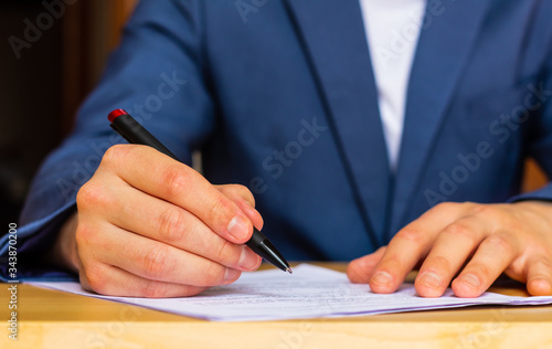 Photographie Close-up of the hands of a well-dressed man in a blue suit and white shirt signing a document on a desk