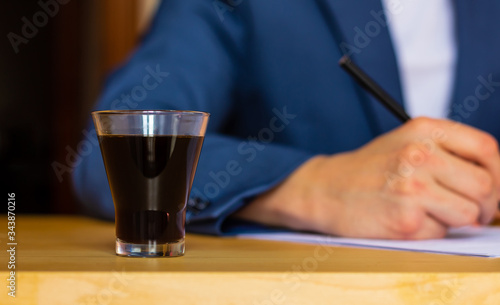 Close-up of a glass of coffee on the table of a well-dressed man in a blue suit and a white shirt writing or signing a blueprint at home or office Poster Mural XXL