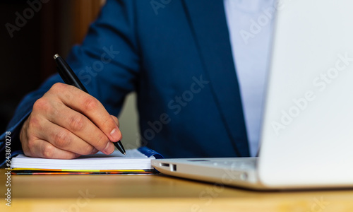 Close-up of the hands of a well-dressed man in a blue suit and a white shirt writing in an agenda or a notebook, with a laptop on a desk Tableau sur Toile