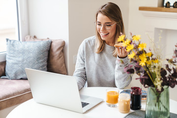 Sticker - Photo of cheerful woman in eyeglasses eating toasts while using laptop